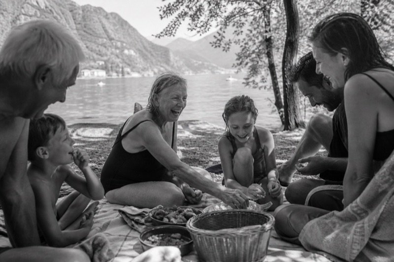Children, parents, and grandparents laughing together during a lakeside picnic after a swim.