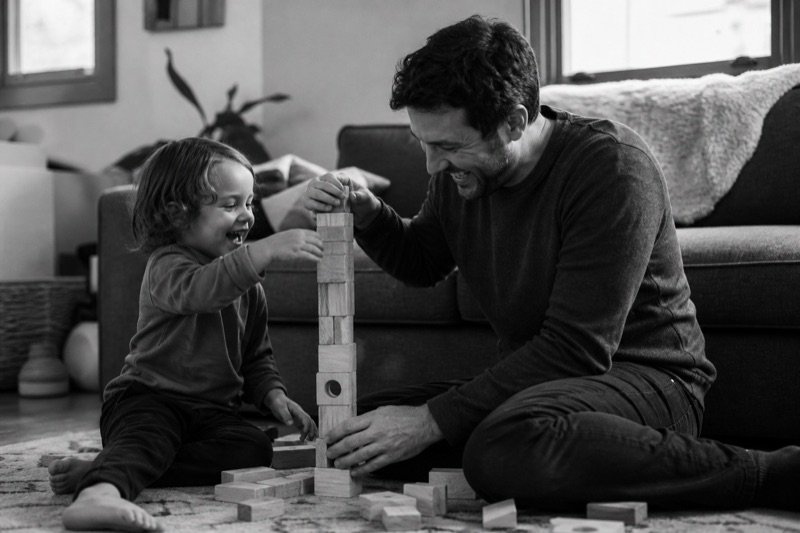 A parent and young child laughing while building a tower from wooden blocks.