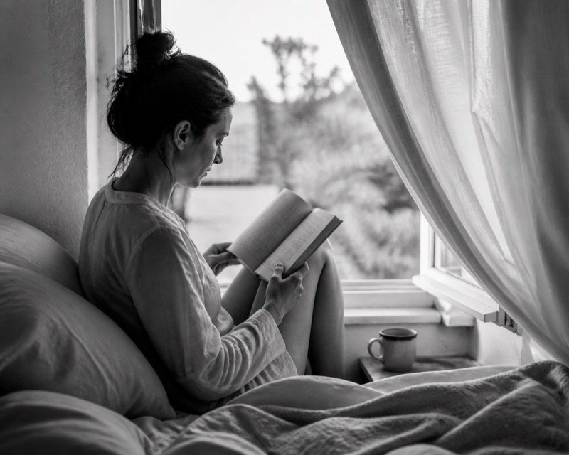 A person reading a paperback beside an open window in the early morning.